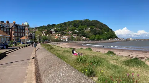 BBC The seafront at Minehead with the Esplanade running behind sand dunes and the beach on a sunny day with the town centre and cliffs in the background