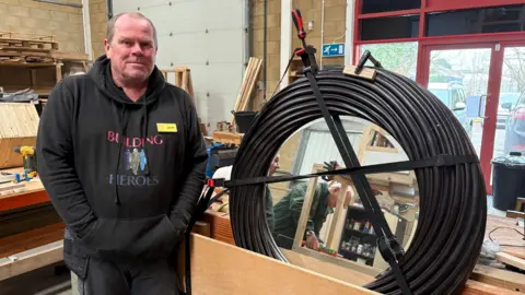 A middle aged man with short, greying hair and a high hairline stands next to a mirror, which he has refurbished with a black, cable-like frame in a workshop. He is wearing a hoodie with colourful silhouettes of soldiers on that says "building heroes", and has his hands in the pockets.