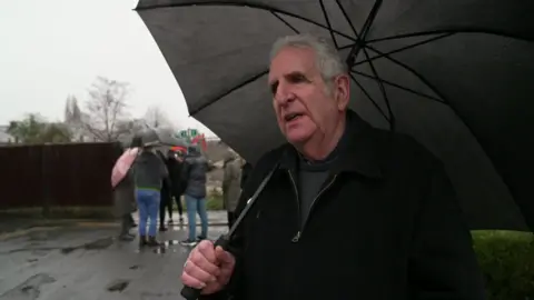John has short grey hair, he holds a black umbrella and wears a black jacket over a dark jumper.