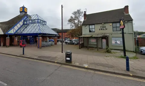 Google An 80s supermarket building with glass pyramid structure over the entrance, next to an older detached pub building in grey with boarded up windows and black lettering reading "The Swan" across the front.