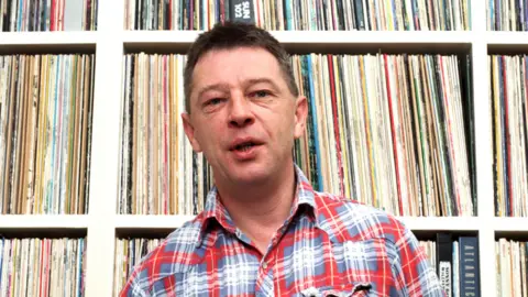 Getty Images Andy Kershaw in a checkered shirt looking at the camera while in front of his record collection