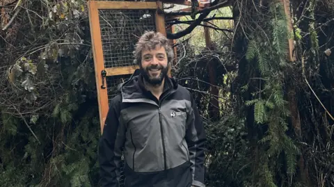 Ed Parr-Ferris stands in front of a large enclosure in woodland. He is wearing a grey and black Wildlife Trust jacket and is smiling at the camera. He has dark brown hair and a beard. 