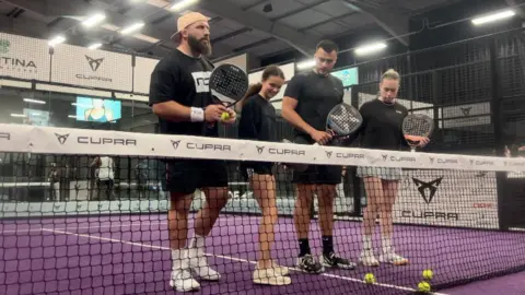 Four people are standing on a purple indoor padel tennis court, holding rackets and wearing sports attire. They are positioned behind the net, which has 'CUPRA' branding on it. The background shows a large indoor facility with additional courts and signage. Tennis balls are scattered near the net.