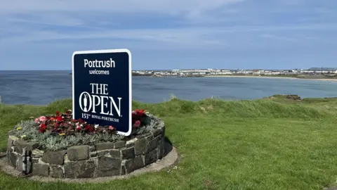 A sign which reads "Portrush welcomes The Open" is placed on a grassy hilltop overlooking the water with Portrush in the background. There are blue skies and the sign is placed in a rockery with flowers.