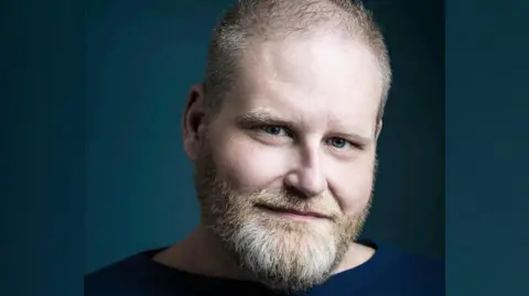 The Seagull Theatre A headshot of John Hales. He is wearing a navy jumper and smiling into the camera. He has a beard. 