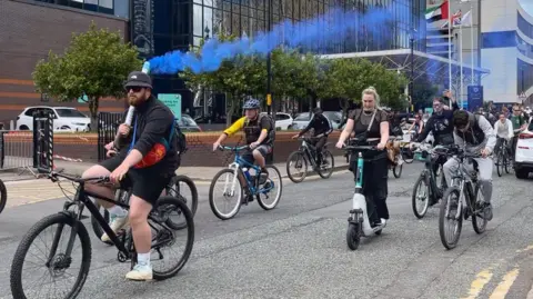 A man is wearing a black top and shorts and is cycling on a bike. He is holding a smoke flare that is letting out blue smoke into the air. People are following him on bikes and scooters