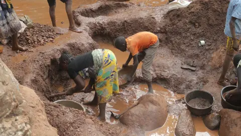 Edwin Naitale / BBC Two women - one in a yellow wrap skirt and dark top, the other in an orange T-shit and three-quarter length trousers - stand in muddy water with bowls as the search for gold.