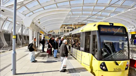 Passengers wait at a platform to get on a Bee Network yellow coloured tram.