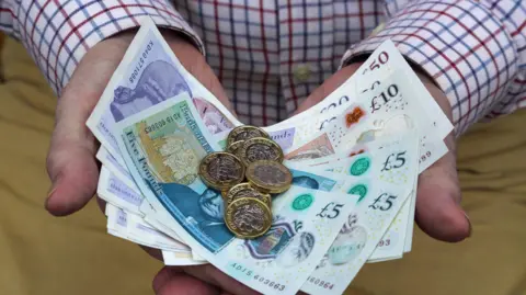 Getty Images Person stood with pound coins and £5 notes in their hands.