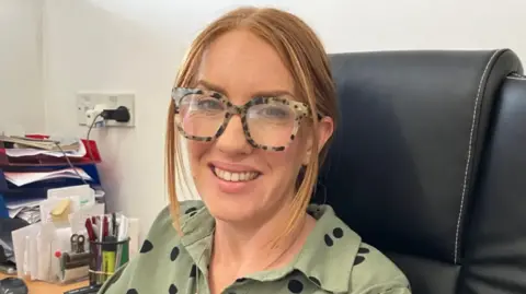 Guy Campbell/BBC Stacey Lock sitting at her desk, smiling at the camera. She is wearing leopard-skin print glasses and a green top with black dots on it. 