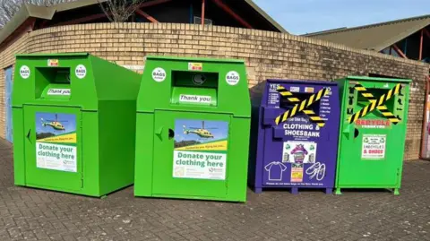 Image of four clothes and shoes recycling banks, lined up against a brick wall. In the background can be seen the roof of a building.
