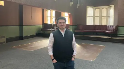 Grace Wood/BBC A man with short brown hair and goatee stands in an empty room with decorative wallpaper and red leather sofas in the background