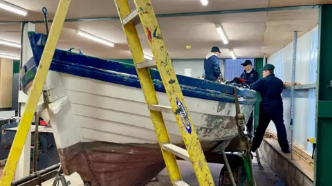 Three young men in blue overalls working on a sailing boat within a workshop, with a ladder in front