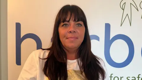 BBC Danielle Chadwick is a woman with long dark brown hair and a straight fringe. She is wearing a white and gold t-shift and is smiling at the camera. Part of the Harbour logo on the wall behind her.