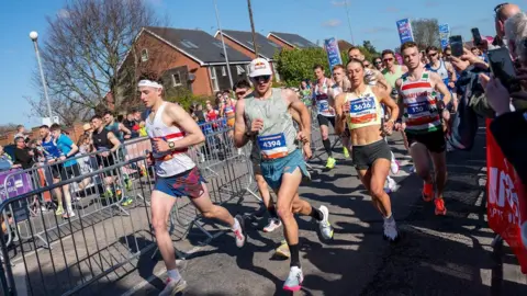 Male and female runners in running shorts and vests are running down a street with houses in the background and members of the public on either side cheering them on from behind metal barriers.