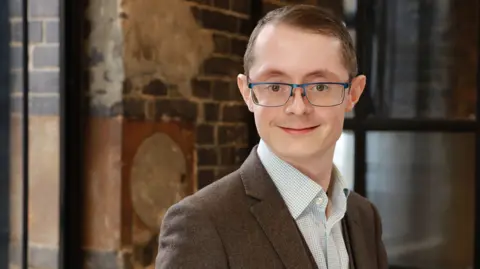 Countryside Alliance David looks into the camera and smiles. He has blue framed glasses and is wearing a brown jacket and light blue checked shirt. Behind him is a bare brick wall and a window. 