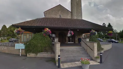 A crematorium, which has a tower and sloping roofs with hanging baskets full of bright flowers, and two green hedges either side of stairs leading to the doors. The sky above is gloomy and grey.