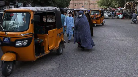 A woman covered with a long robe from head to toe, with her face covered by a medical mask, walks down the street near a tuktuk. Men can be seen in the background