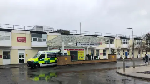 The main entrance to Frimley Park hospital which has a glass canopy over the doors and is a two storey building rendered in a pale yellow.