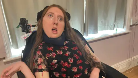 A person sitting in a powered wheelchair indoors, wearing a black dress with a red and pink floral pattern. The wheelchair is positioned in front of closed curtains.
