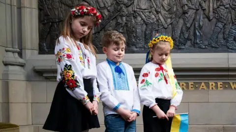 Three young Ukrainian children look down at poppy wreaths at a war memorial. They are wearing Ukrainian national dress. Two girls are wearing flower garlands in their hair and one is holding a small Ukrainian flag  