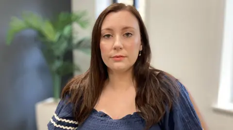 A woman with long brown hair is looking at the camera. She is wearing a blue top and behind her is a plant.