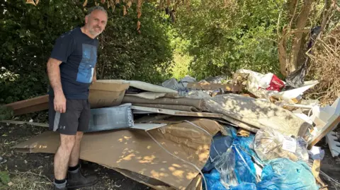 Man stands next to a large pile of rubbish.