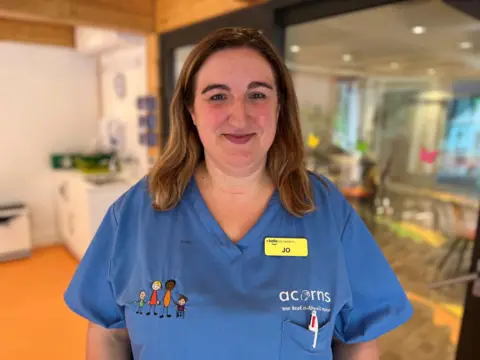 A woman in blue medical scrubs smiles at the camera. She has straight brown hair and a yellow name badge that says "Jo". There is a kitchen behind her.