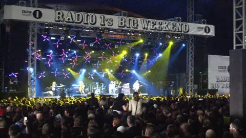 The Radio 1 Big weekend stage has black-and-white branding and crowds of people watching on. There is a band on stage under multi-coloured lights.