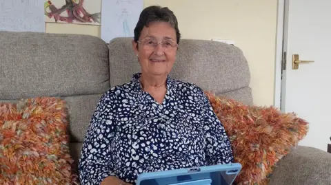 BBC Christine Wallace has short dark hair and glasses. She is wearing a patterned blouse and is sitting on a sofa and using an Ipad.