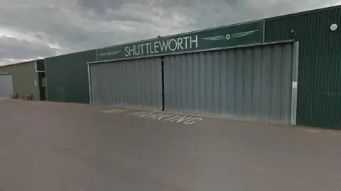 Google The outside of an aircraft hangar at Shuttleworth in Biggleswade. The outside of the building is dark green and there is a sign on top which reads SHUTTLEWORTH in white lettering.