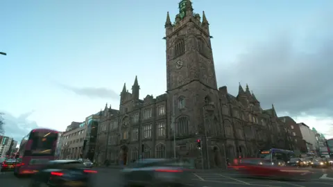 BBC A large church building in Belfast city centre shows, stained glass windows and a bell tower, bearing a gold and black clock. Behind the building is a gloomy, cloudy sky. Traffic is blurred as it passes the camera. 