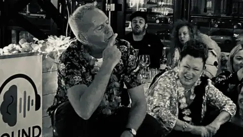 Tom Breen Fatboy Slim, AKA Norman Cook, wearing a floral short-sleeve shirt, sits with group of people discussing DJ mixing at the Sounds Mind event. It is a black and white photograph.