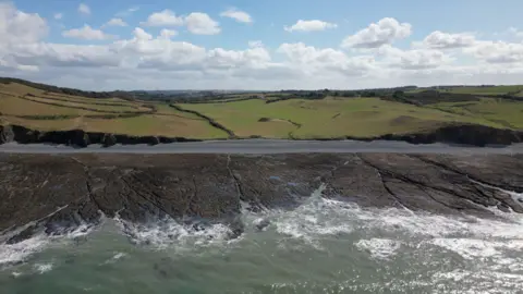 A aerial view of the coastline, showing waves breaking onto a rocky foreshore. there are gently sloping fields beyond