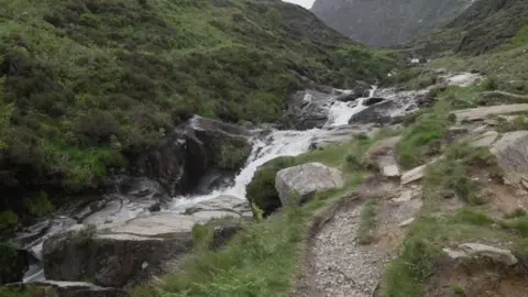 A mountainside with greenery on either side and water running down the middle in a stream with lots of boulders and rocks