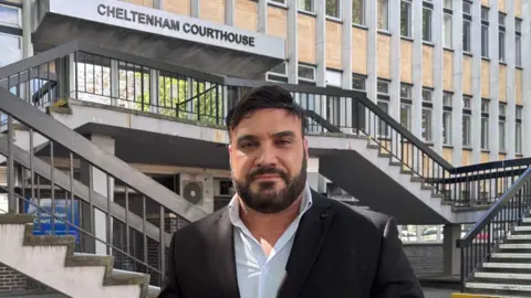 A man in his 40s with short brown hair swept to the side and dark facial hair smiles as he stands outside a court. Behind him is a courthouse, two flights of steps and a building with numerous windows.