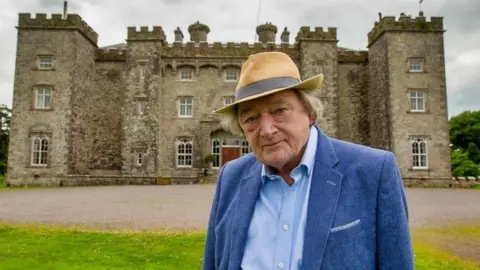 Lord Mount Charles wearing a blue blazer and shirt, he is also wearing a tan hat. He has grey hair. Behind him is a castle