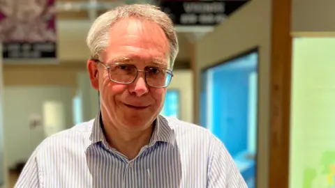 Shaun Whitmore/BBC Dr Tim Pestell standing in a museum. He is looking directly at the camera and smiling and he is wearing a white and blue striped shirt and a pair of glasses.