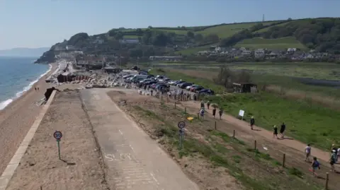 A drone shot of a group of people walking along a part of the A379 between Torcross and Slapton on a clear sunny day. There are lots of cars parked. The sea is visible and grass land in the background.