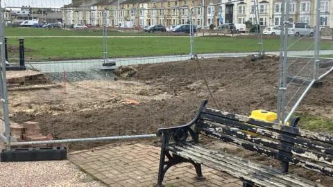 GUY CAMPBELL/BBC In the foreground, a weathered, black bench sits on brick paving. There is metal fencing behind it and beyond the fencing, the ground has been dug up. In the distance, the other side of a green, there are terraced houses. 