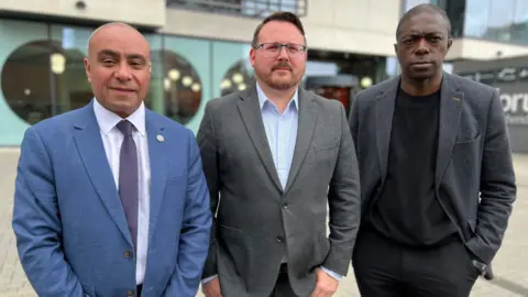 Stuart Woodward/BBC David Burton-Sampson, Daniel Cowan and Bayo Alaba stand next to eachother in front of The Forum building. David wears a blue suit with light blue shirt and a purple tie. Daniel wears a grey suit with open-neck light blue shirt. Bayo wears a dark grey jacket over a black shirt.