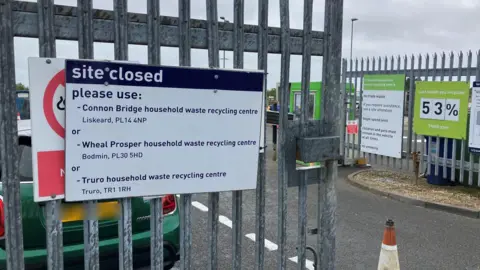 BBC White and navy blue sign on the entrance to the St Austell Household Waste and Recycling Centre site says "site closed" and tells people to use three alternative locations. A green Mini Cooper is parked the other side of the main gate.