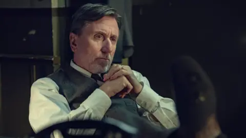 Netflix Tim Roth in a white shirt and grey waistcoat, his hands clasped and looking pensive in a scene from the film