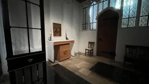 Shaun Whitmore/BBC A small, dimly lit chapel interior with a simple design. At the center is a wooden altar featuring a carved cross symbol on its front panel. Two tall white candles in black holders stand on either side of the altar, and above it hangs a framed religious painting depicting several figures. 