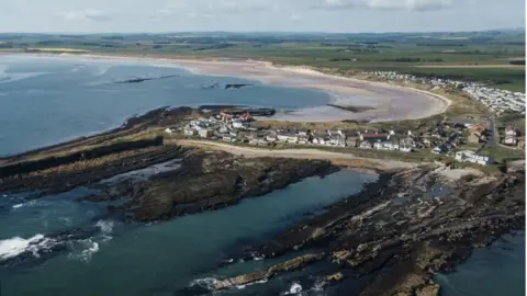 Solstock  An aerial view of Beadnell showing a row of houses along the cliff and the sweep of the bay behind 