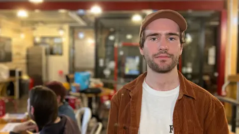 Sam is standing indoors in front of a café-style background. He is wearing a white T-shirt under a rust-colored corduroy jacket and a cap. Behind them, there are tables with people seated and a wall with posters on the left side. The space has warm lighting and industrial-style décor.