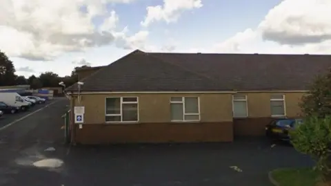 A single storey brown building with white rimmed windows. There are cars parked to the left of it.