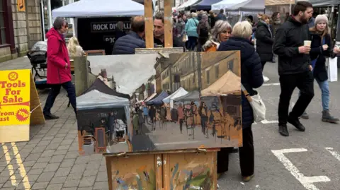 Warminster Town Council Image shows people walking past market stalls, two people are seen holding cups of coffee, others are chatting and some are walking in the opposite direction. In front of them is a painting which of a busy market stall. 