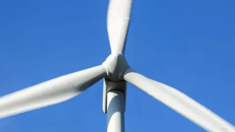 Getty Images A close up of the top of a wind turbine. In shot is its three large blades and hub. The turbine is pictured against a blue sky.