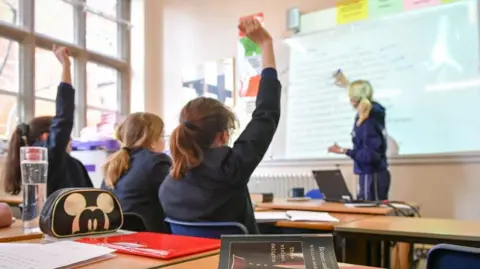 PA Media The image shows a classroom during a lesson in a secondary school setting.
Several students are seated at desks in the foreground and middle ground. At least three students have one arm raised. The students are wearing dark school uniforms. At the front of the room, a teacher stands near an interactive whiteboard.